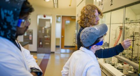 2 students watching another student write an equation in front of a chemistry instrument