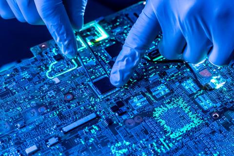 Closeup photo shows person wearing blue gloves using tweezers to work on a semiconductor circuit board